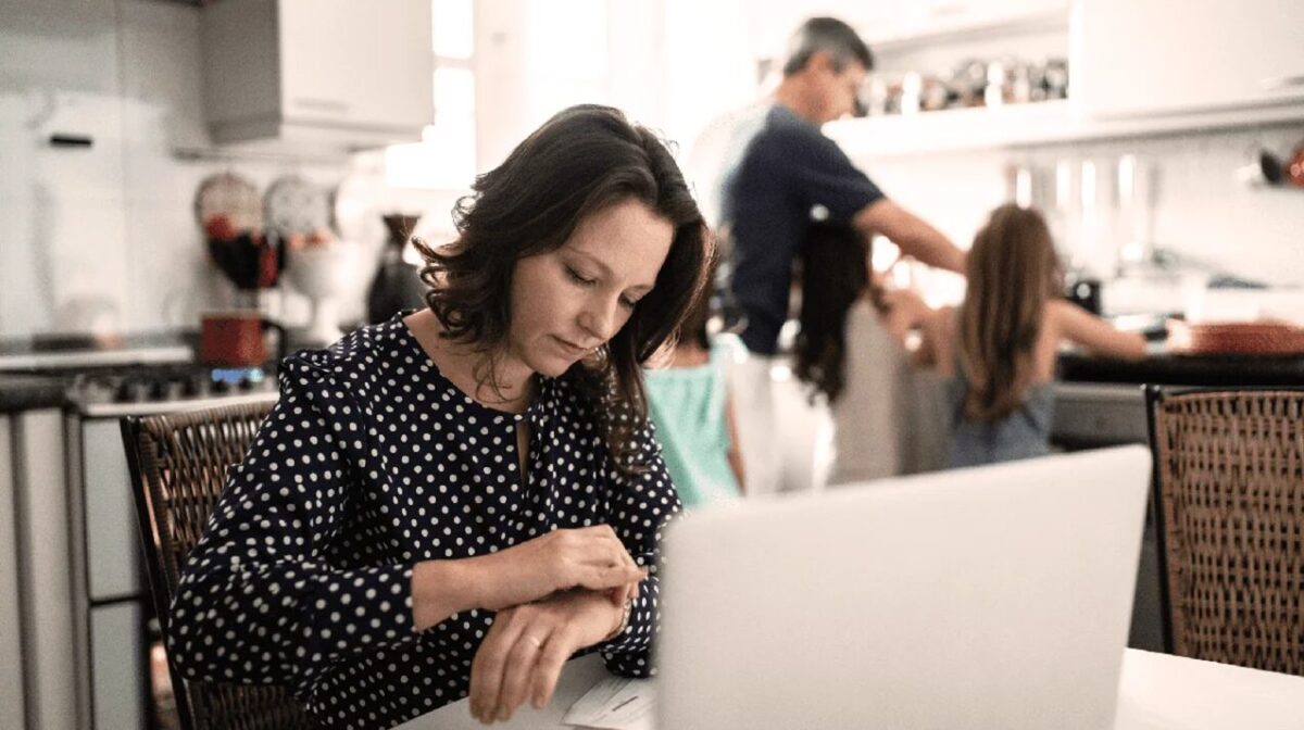 Busy woman checking her watch in front of a laptop in the kitchen with her family in the background.