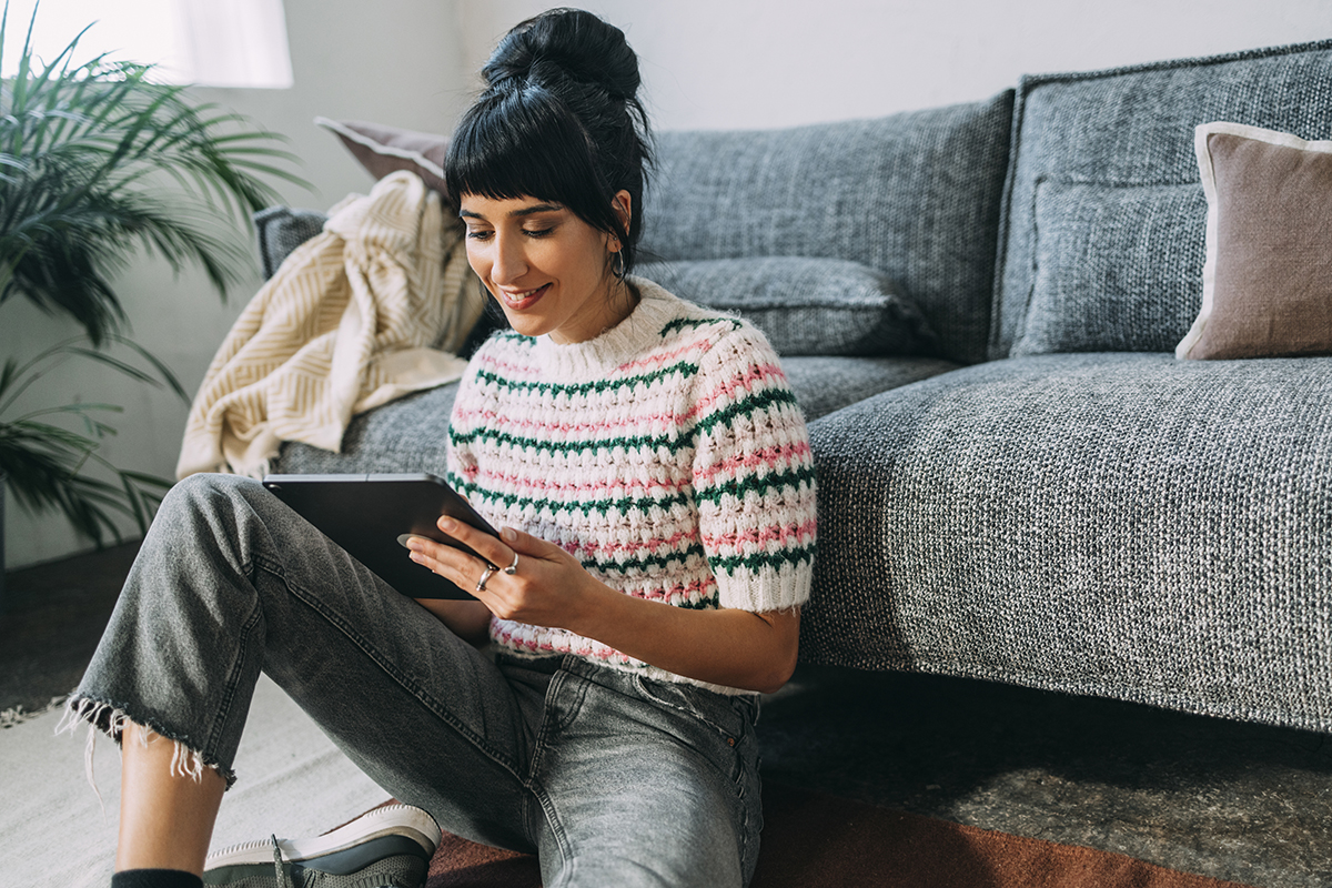 Smiling woman sitting on the floor next to her couch while using a tablet device.