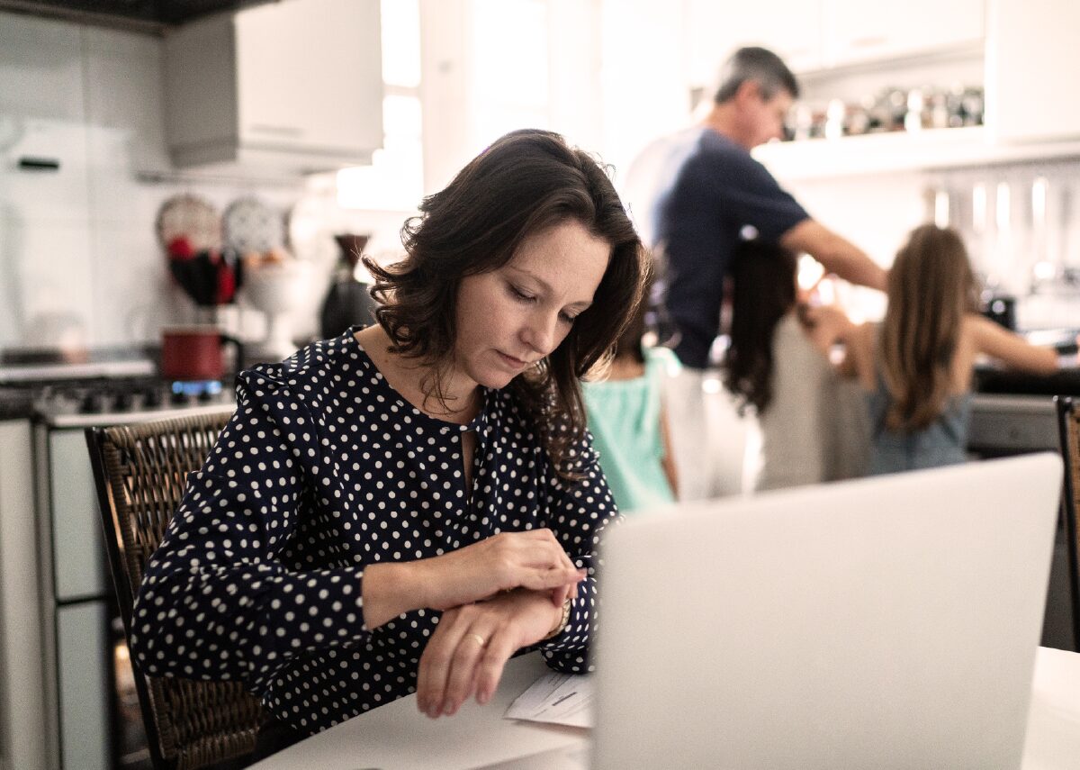 Worried female employee checking the time on her watch while working on her laptop from home, while her husband and daughter prepare a meal in the background.